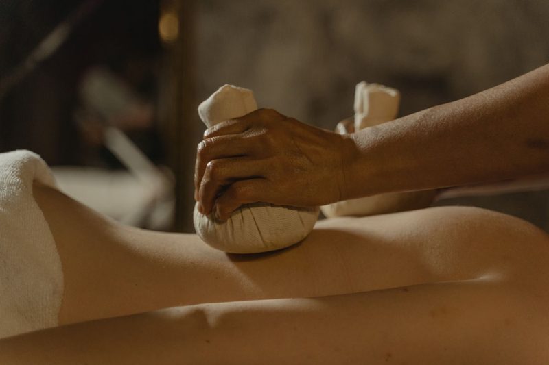 A therapist's hands working warm herbal compresses along a client's back